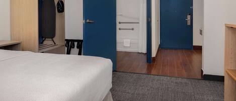 A hotel room with a bed, a wardrobe, a bathroom with a shower, and a door leading to another room.