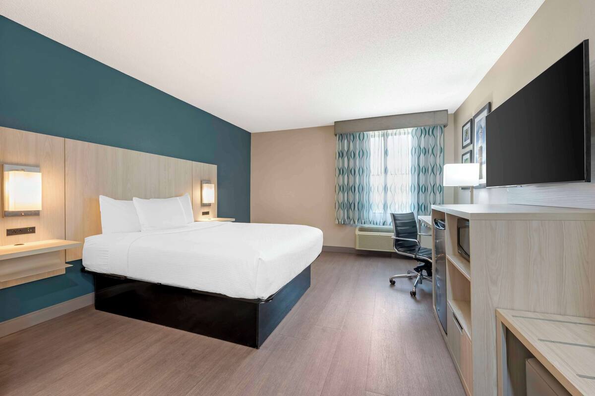 A modern hotel room with a bed, bedside tables, a desk with a chair, a flat-screen TV, and a window with curtains.