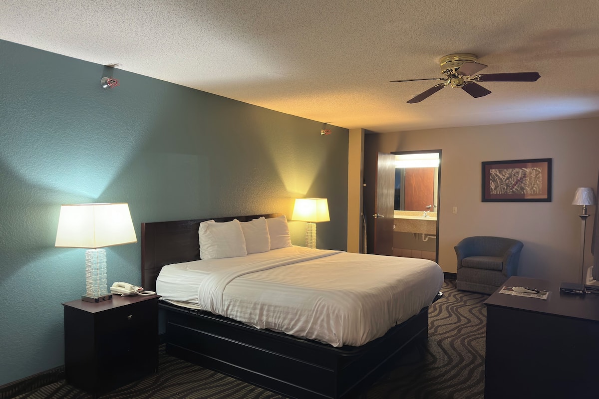 Une chambre d’hôtel comprenant un lit, deux lampes de chevet, un ventilateur de plafond, un bureau avec une lampe, une chaise et un miroir dans la salle de bain.