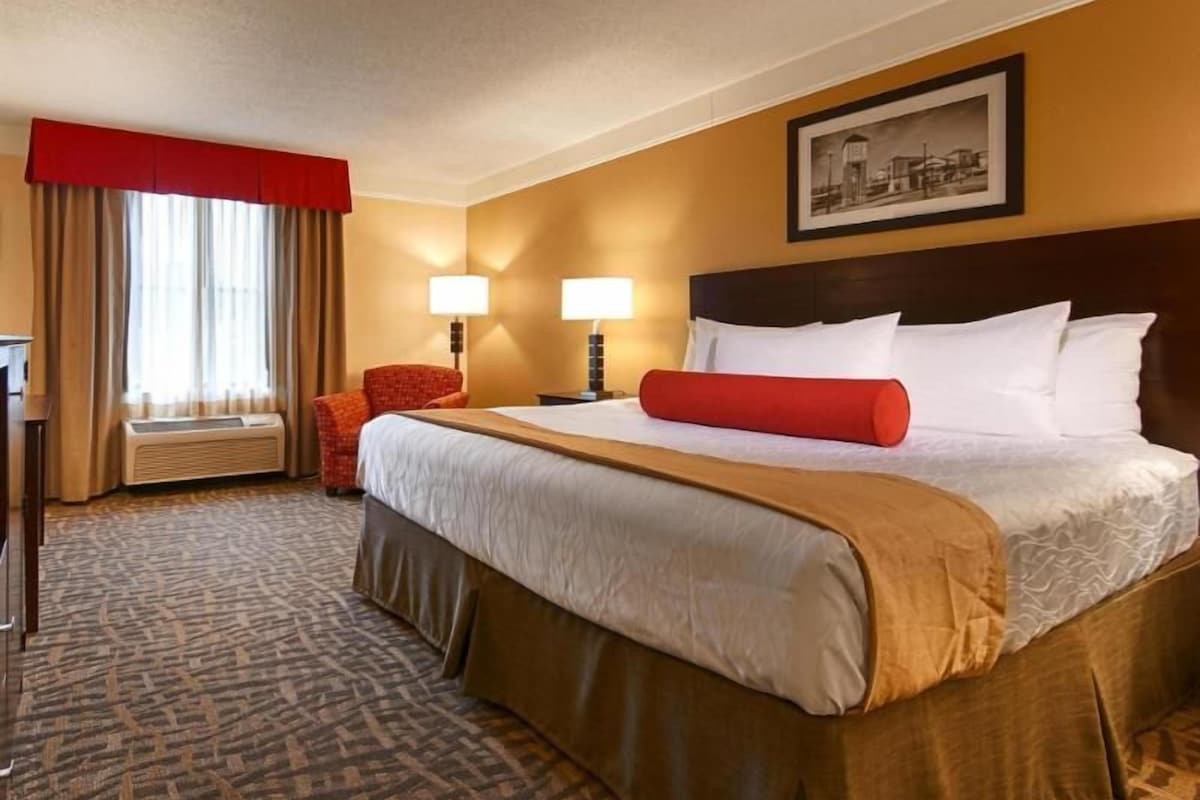 Ein Hotelzimmer mit einem großen Bett, zwei Nachttischlampen, einem roten Zierkissen, einem roten Sessel, einem Schreibtisch und einem Fenster mit Vorhängen.