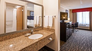 A hotel bathroom with a granite countertop sink, a large mirror, a shower, and a desk with a chair.