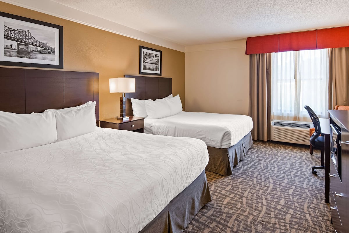 A hotel room with two beds, a desk, a chair, a window with curtains, and a wall-mounted air conditioner.