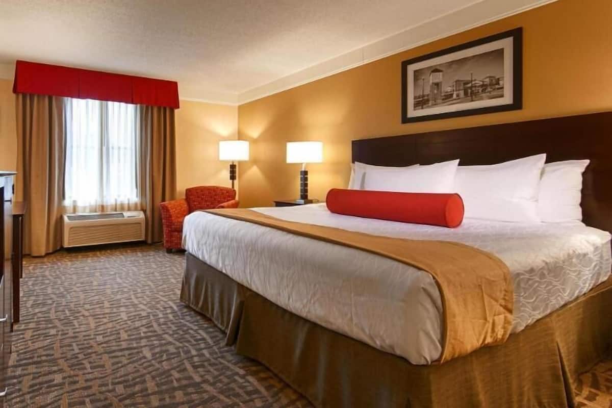 A hotel room with a large bed, two bedside lamps, a red accent pillow, a red chair, a desk, and a window with curtains.