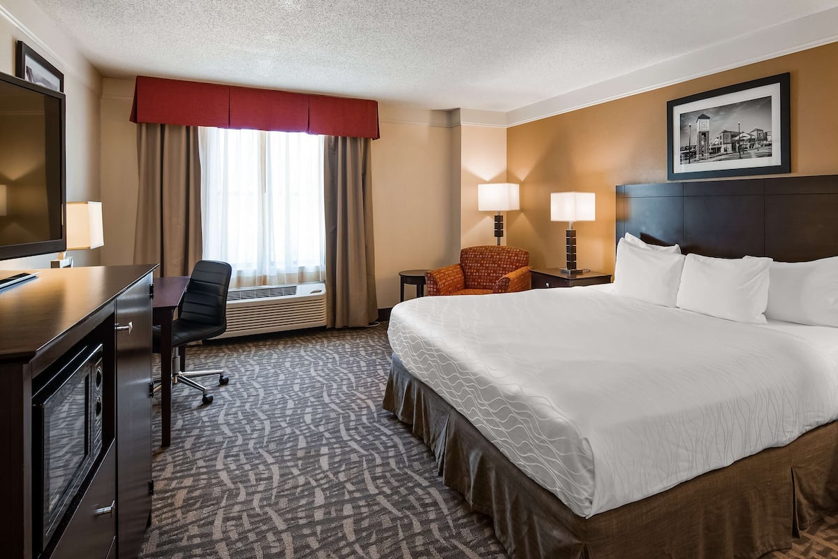 A hotel room with a large bed, a desk with a chair, a small table with a lamp, and a picture on the wall.