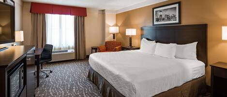 A hotel room with a large bed, a desk with a chair, a small table with a lamp, and a picture on the wall.