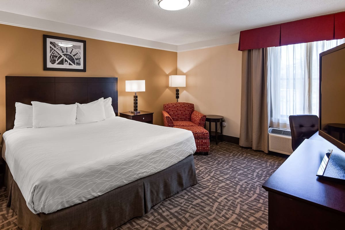 A hotel room with a large bed, two bedside tables with lamps, a red armchair, a desk with a chair, and a large window with curtains.