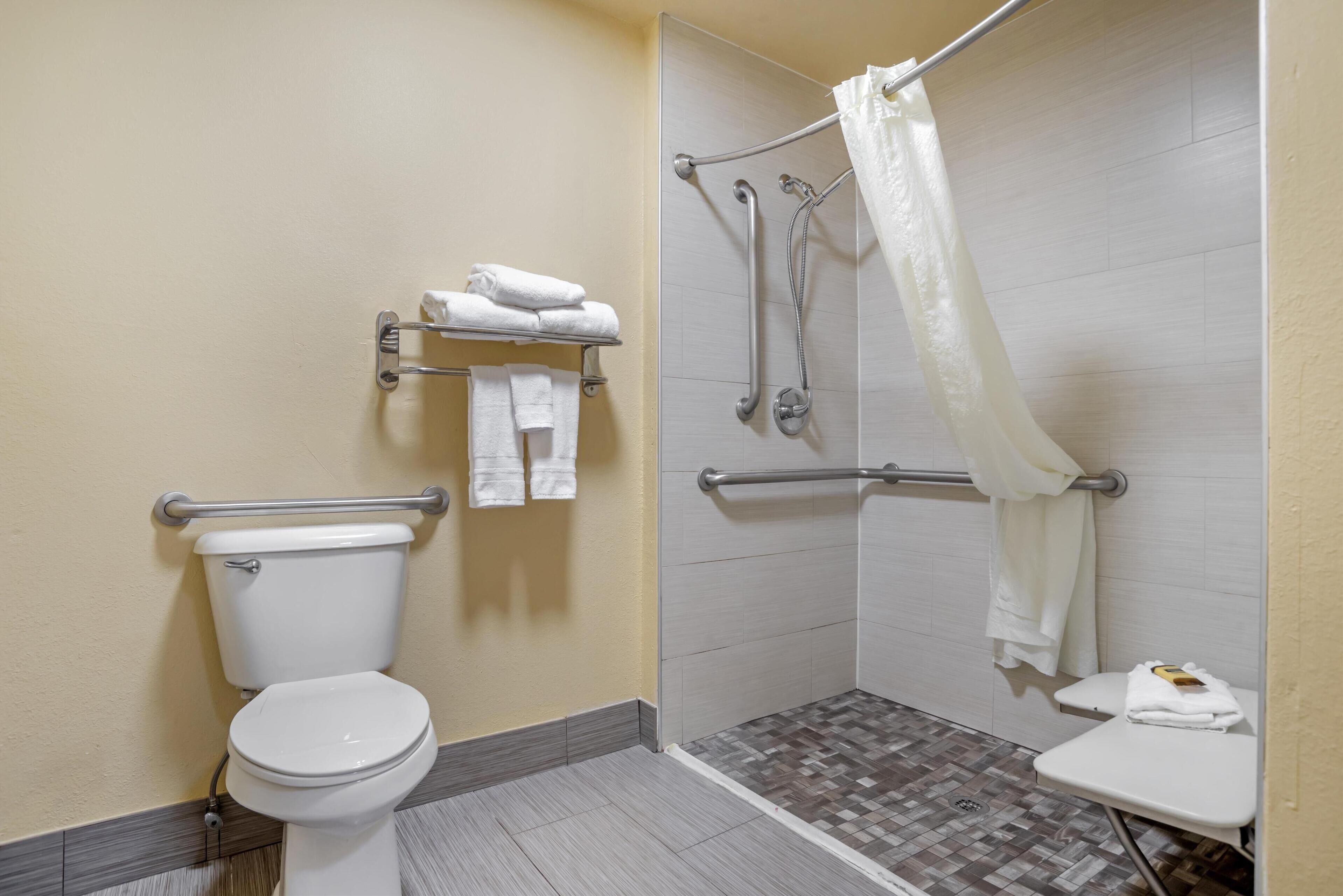 A wheelchair-accessible bathroom with a toilet, grab bars, a shower with a white curtain, and a small table with a towel.