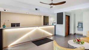 Zona de recepción de hotel con diseño moderno, que incluye un mostrador de recepción, un ventilador de techo y un área de descanso con una mesa y sillas.