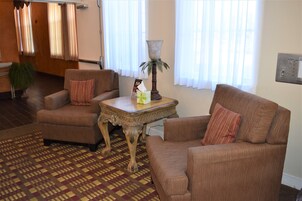 Un salón con dos sillones marrones, un tapete estampado, una mesita con lámpara y un ventanal con cortinas.