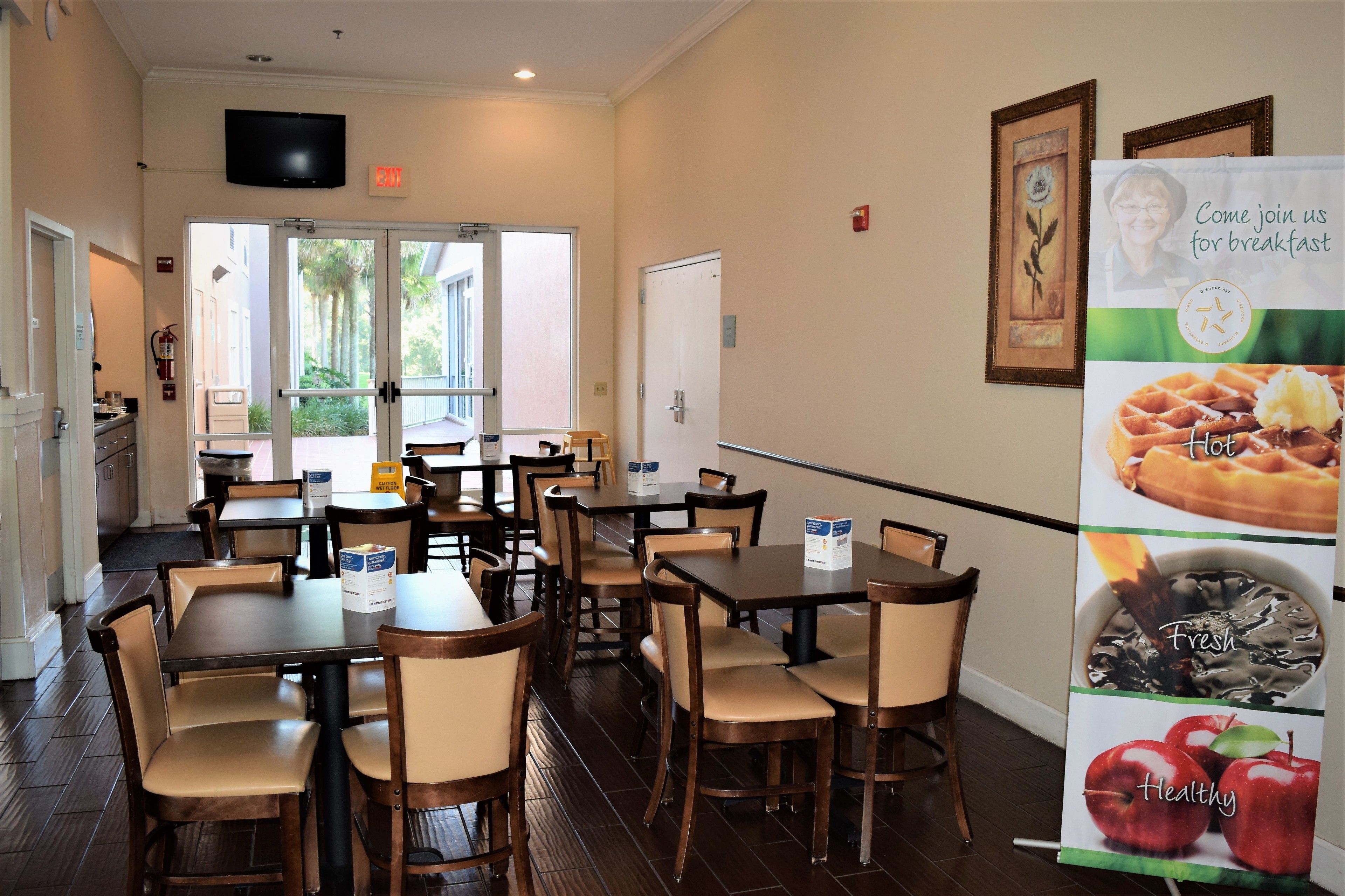 A dining area with tables and chairs, a kitchen in the background, and a poster advertising breakfast.