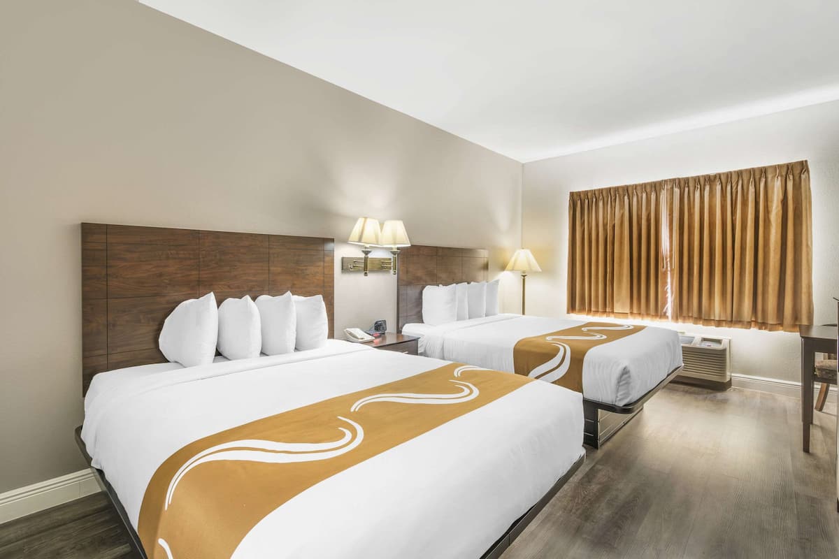 A hotel room with two beds, a wooden headboard, bedside lamps, a window with curtains, and a desk with a chair.