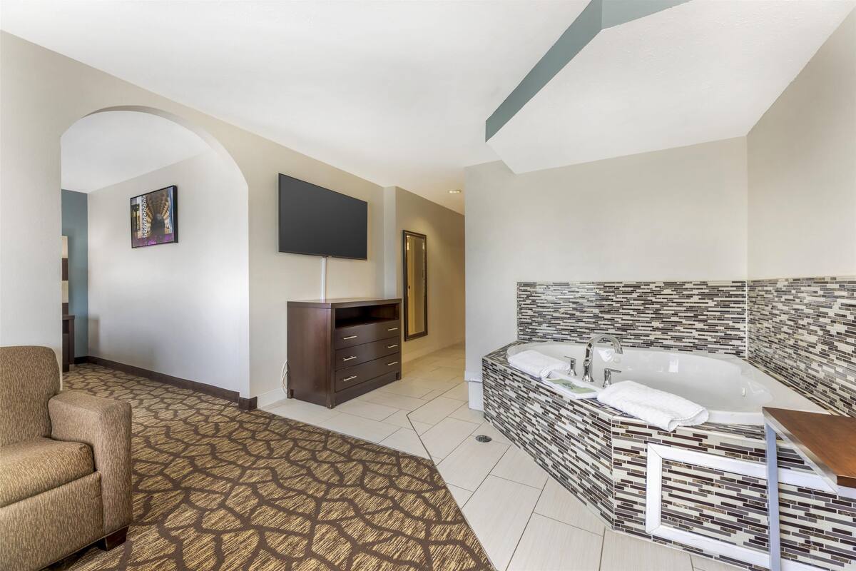 A modern hotel room with a large bathtub, a flat-screen TV, a sofa, and a patterned carpet.