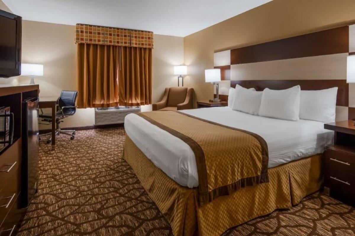 A hotel room with a large bed, a desk with a television, two chairs, a desk lamp, and a window with curtains.