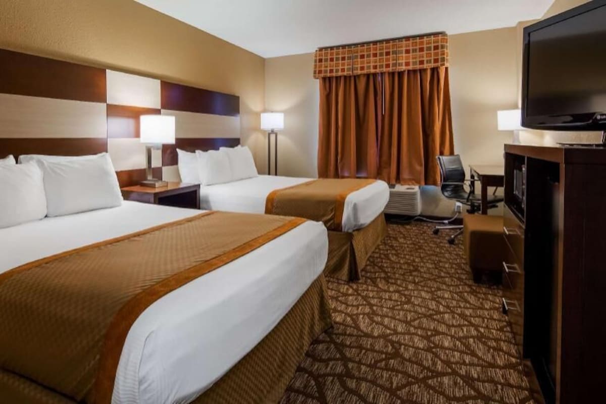 A hotel room with two beds, a television, a desk with a lamp, and a patterned carpet.