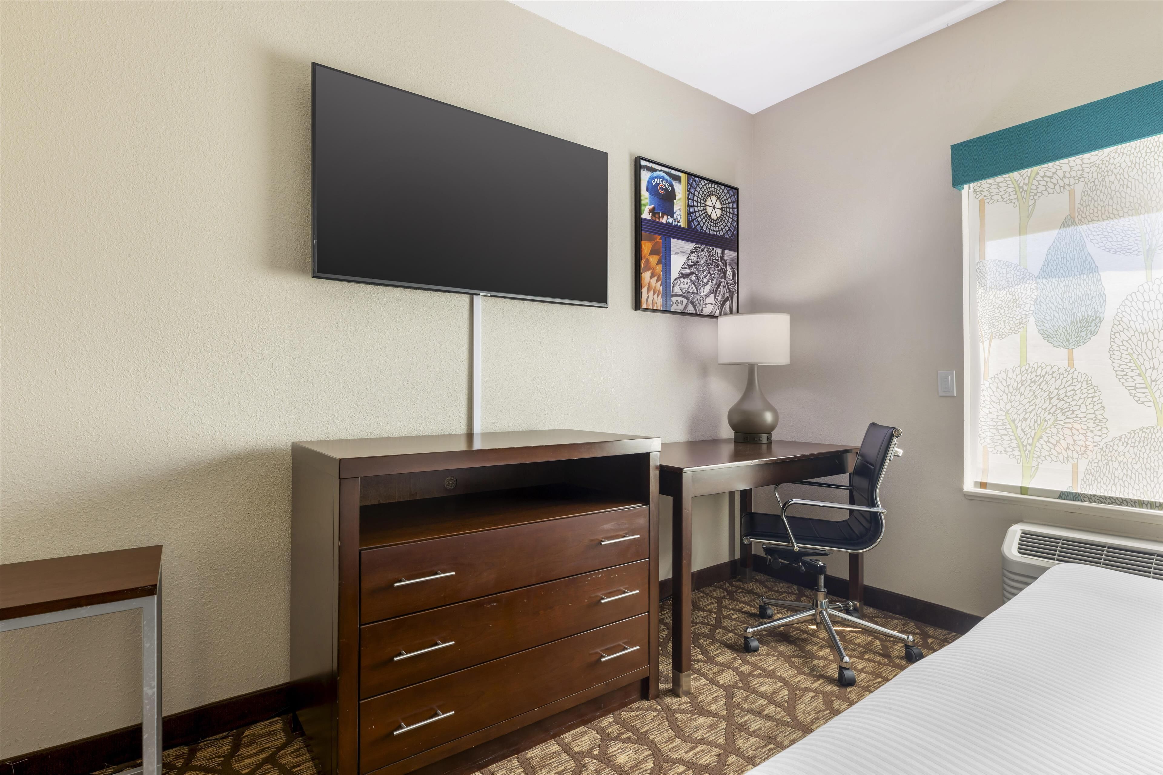 Une chambre d’hôtel équipée d’un téléviseur à écran plat fixé au mur, d’un bureau avec une chaise, d’une commode et d’une fenêtre donnant sur la verdure.