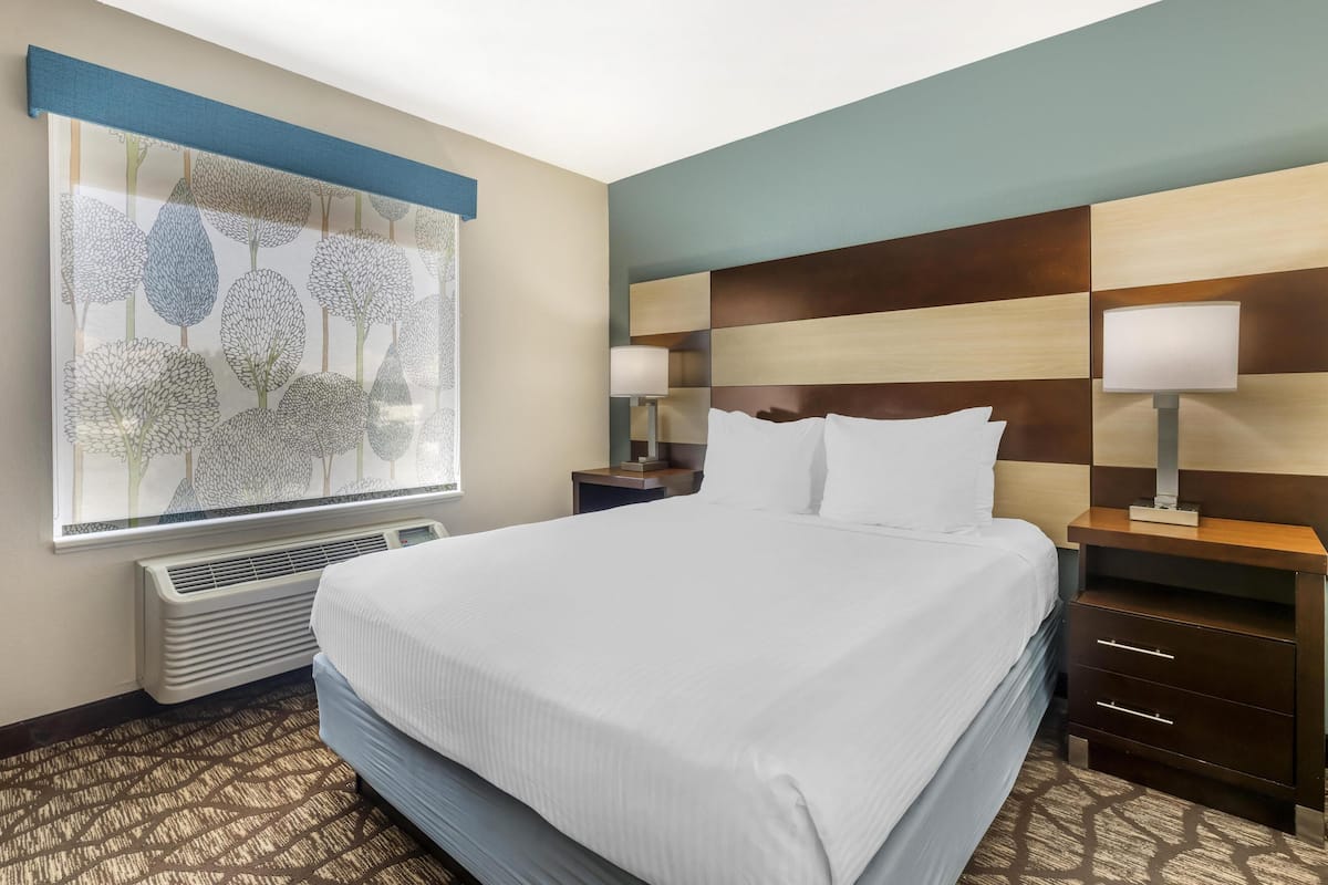 A hotel room with a large bed, a nightstand with a lamp, a wall-mounted air conditioner, and a decorative window blind.