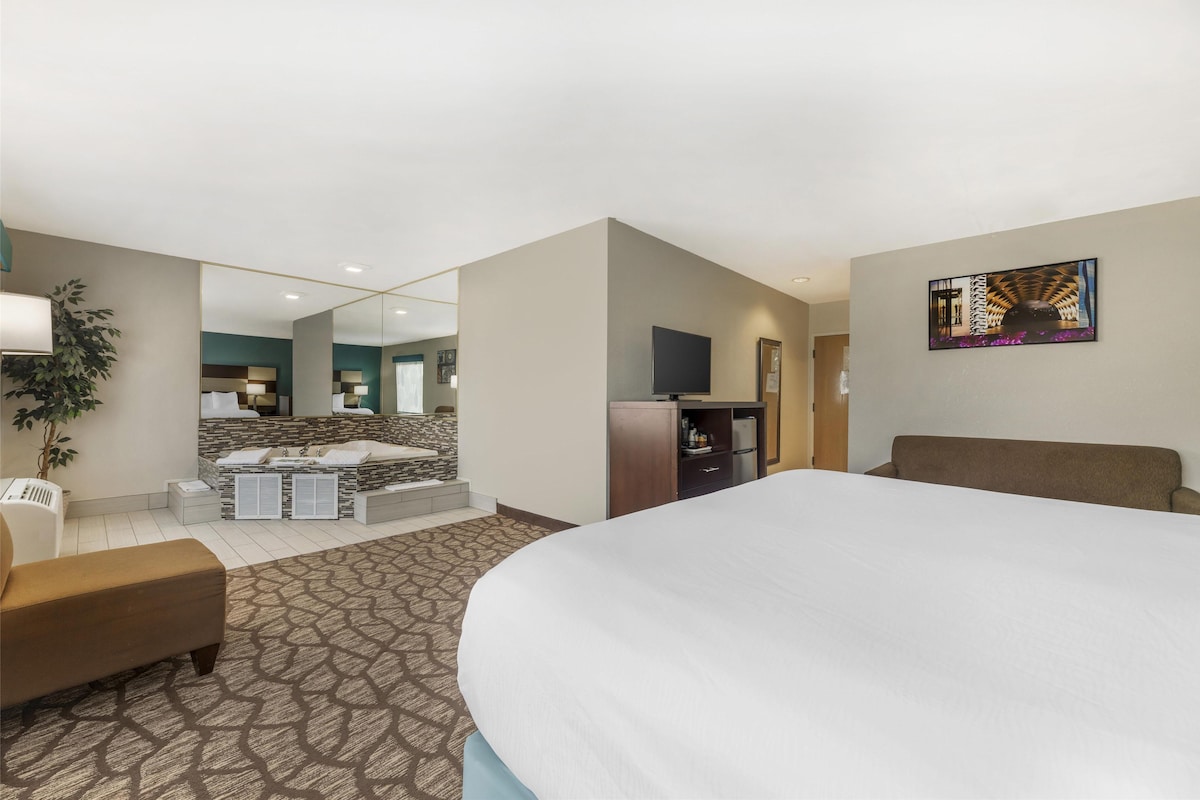 A hotel room with a large bed, a sofa, a TV, a desk, and a bathroom with a shower and a mirror.