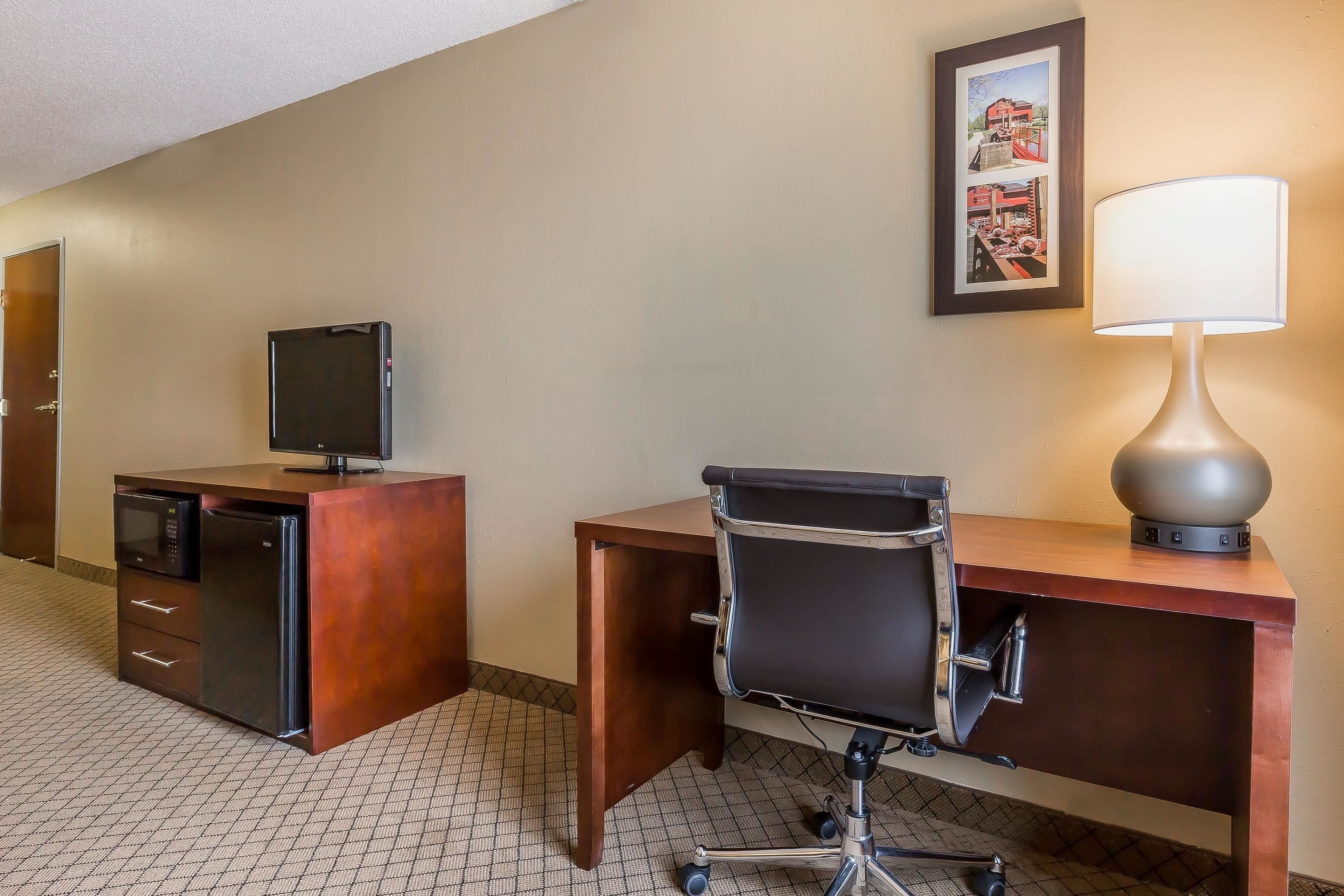 Een hotelkamer met een houten bureau, een zwarte bureaustoel, een televisie, een lamp en een lijst aan de muur.