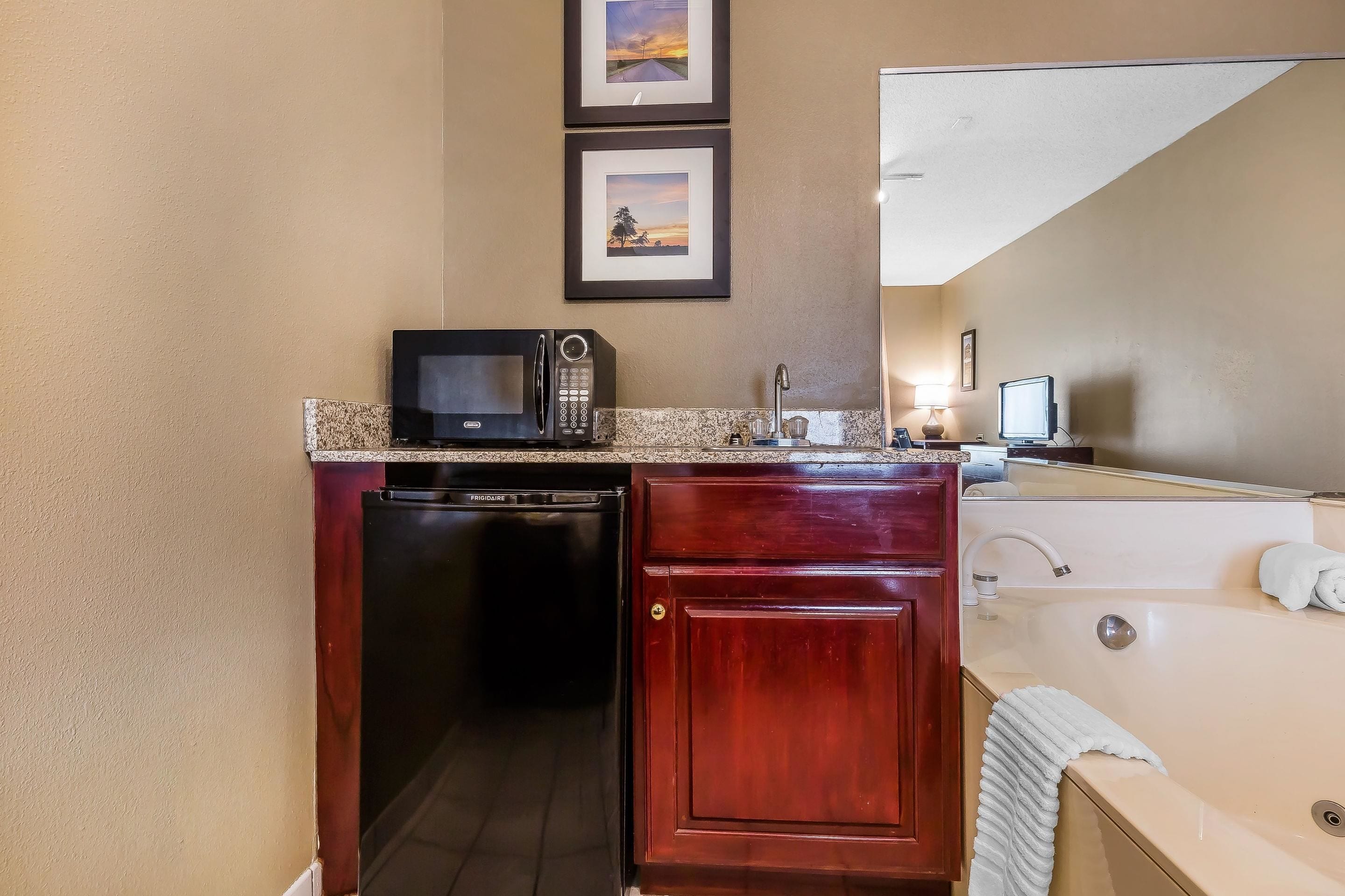 A compact kitchenette with a microwave, dishwasher, and a small refrigerator. There is a sink with a faucet and a mirror above it. A white towel hangs on the edge of the sink.