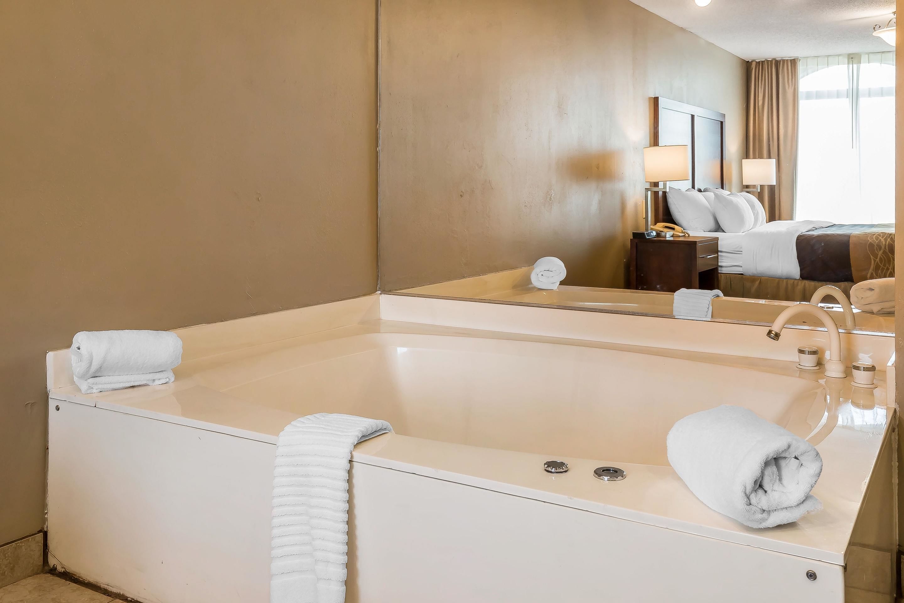 A hotel bathroom with a large bathtub, a mirror, and a bed with a headboard.
