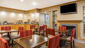 A dining area with wooden tables and red upholstered chairs, a kitchen in the background, and a fireplace with a TV above it.