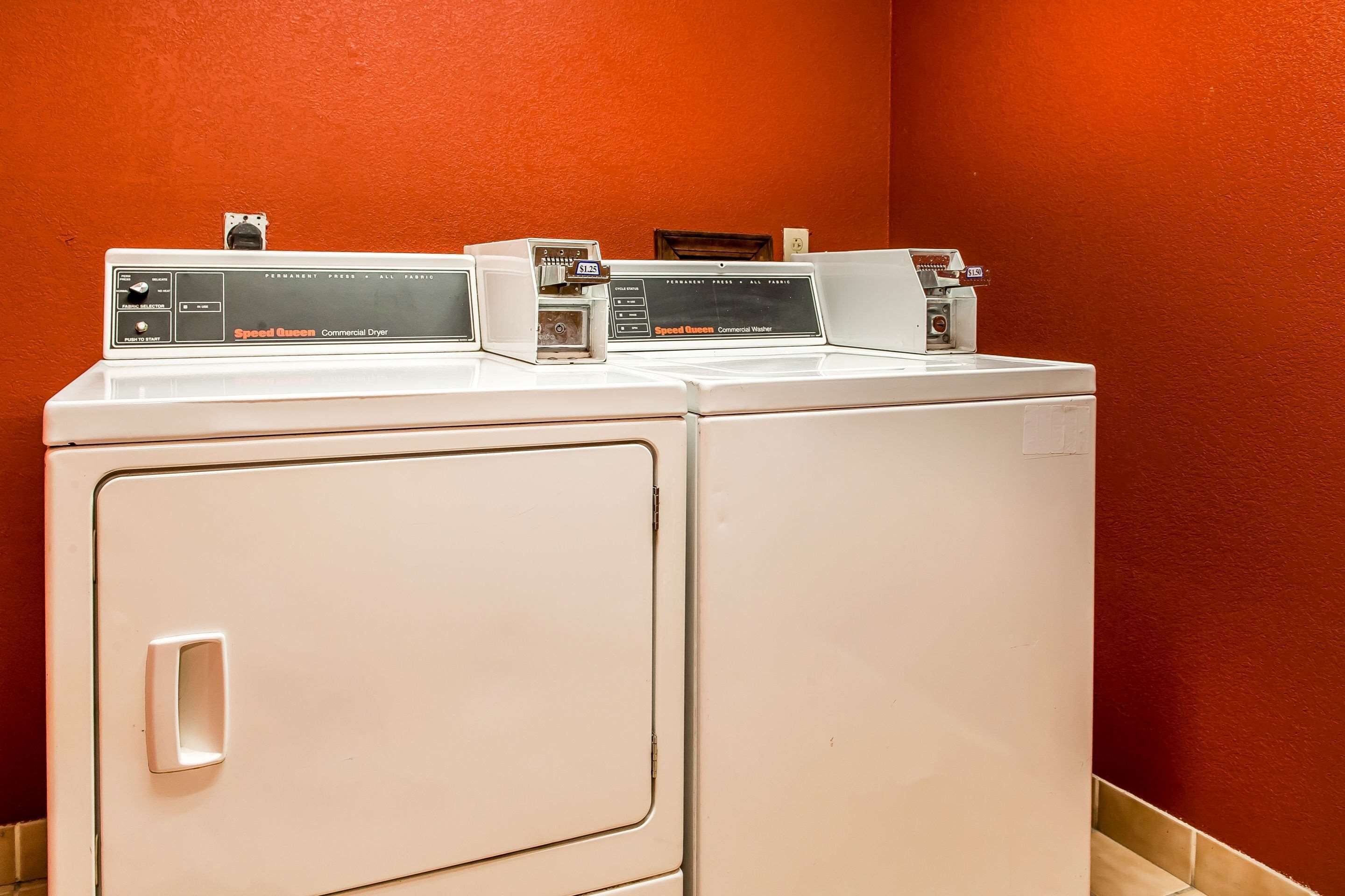 A white commercial dryer with a control panel labeled "Speed Queen Commercial Dryer" in a laundry room with a red wall.