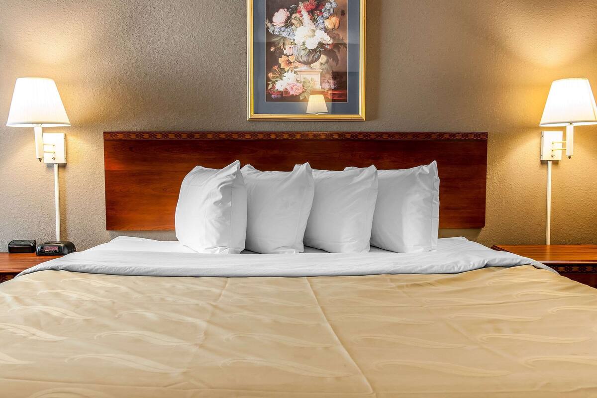 A hotel room with a large bed, white bedding, a wooden headboard, two bedside lamps, and a framed floral artwork on the wall.