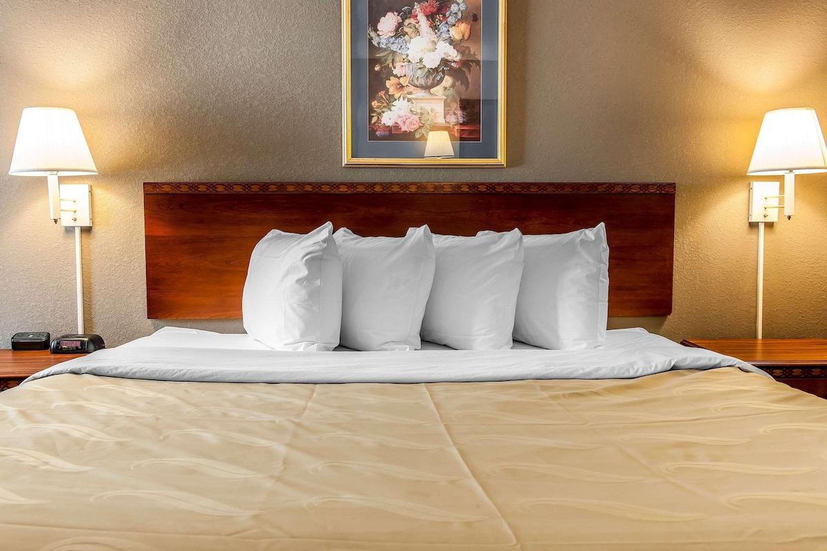 A neatly made bed with white linens and a beige comforter, flanked by two bedside lamps and a framed floral artwork on the wall.