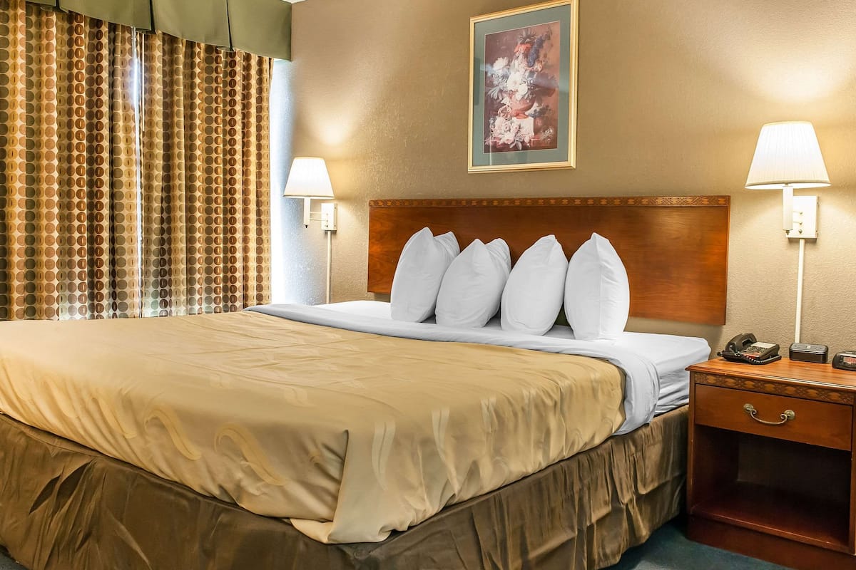 A hotel room with a large bed, a wooden headboard, two bedside lamps, a nightstand with a phone, and a painting on the wall.