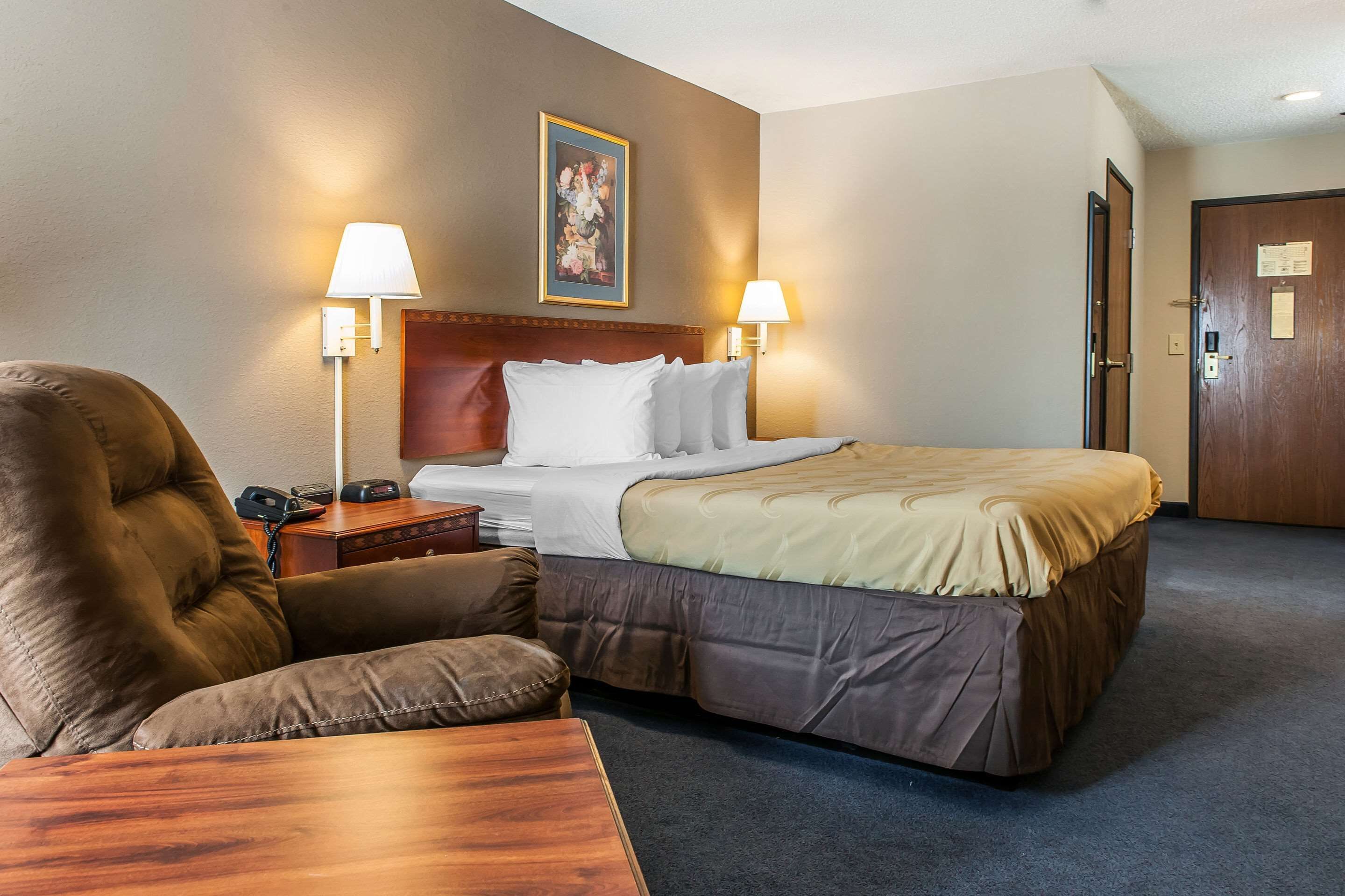 Een hotelkamer met een groot bed, twee bedlampen, een houten nachtkastje, een bruine fauteuil en een houten tafeltje.