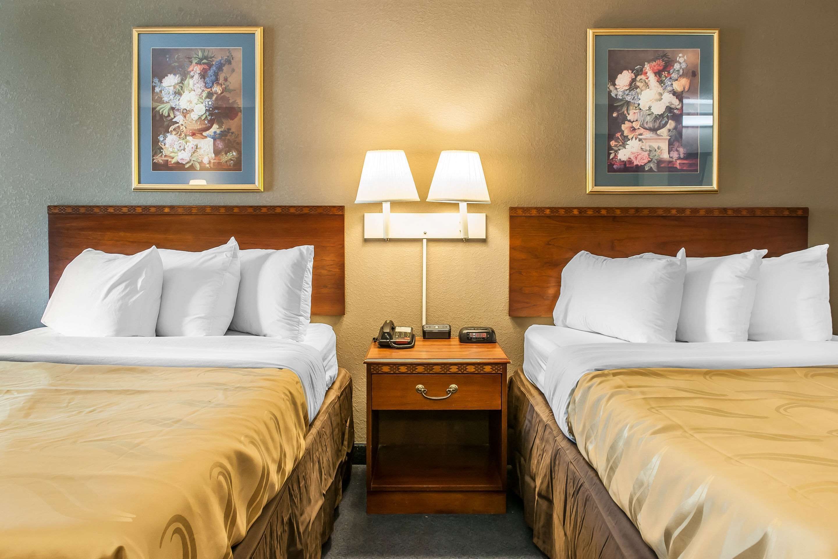 A hotel room with two beds, a nightstand with a lamp, and two framed floral paintings on the wall.