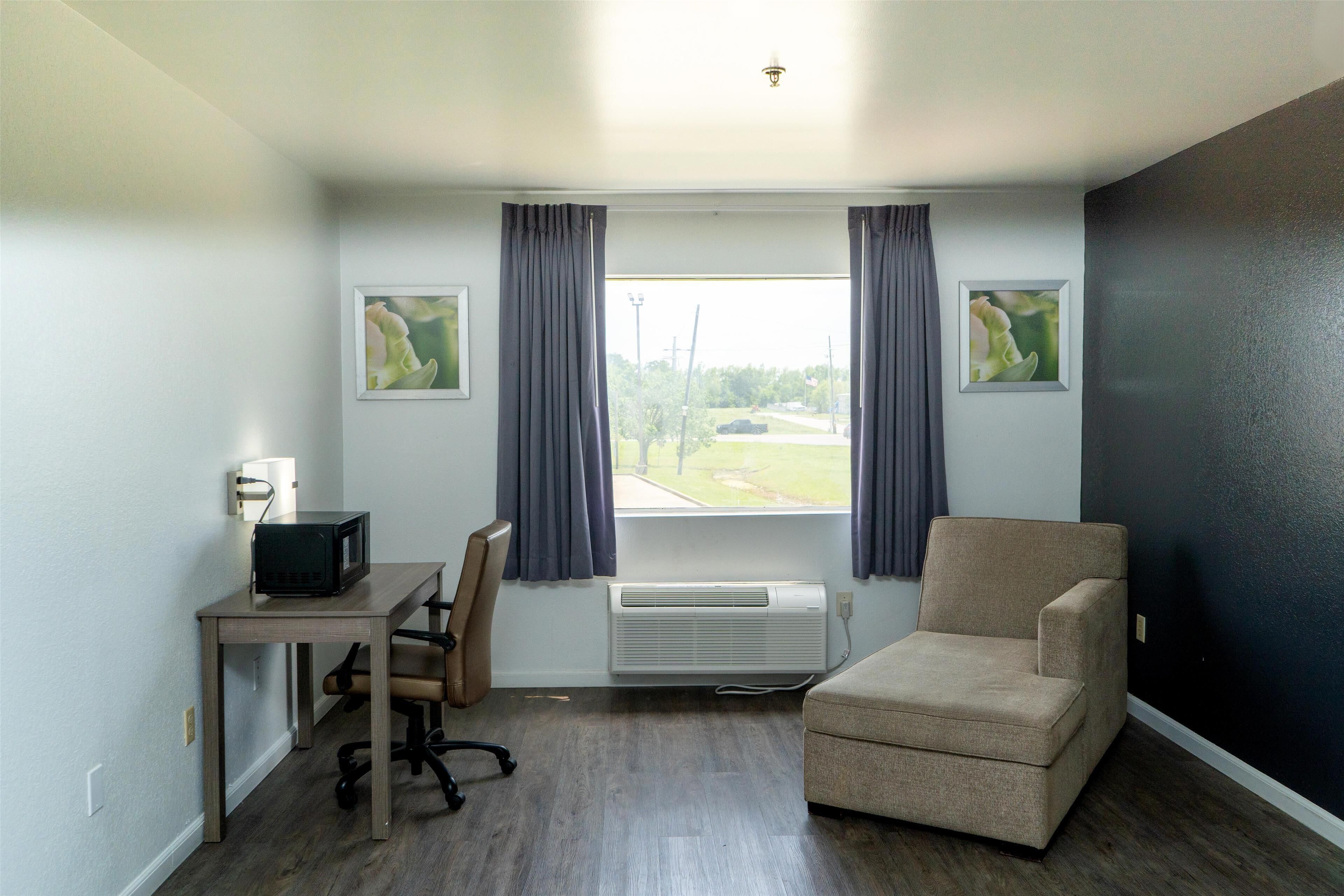 Ein Zimmer mit Schreibtisch, Stuhl und Sesseln, ein Fenster mit Vorhängen und zwei gerahmte Bilder an der Wand.