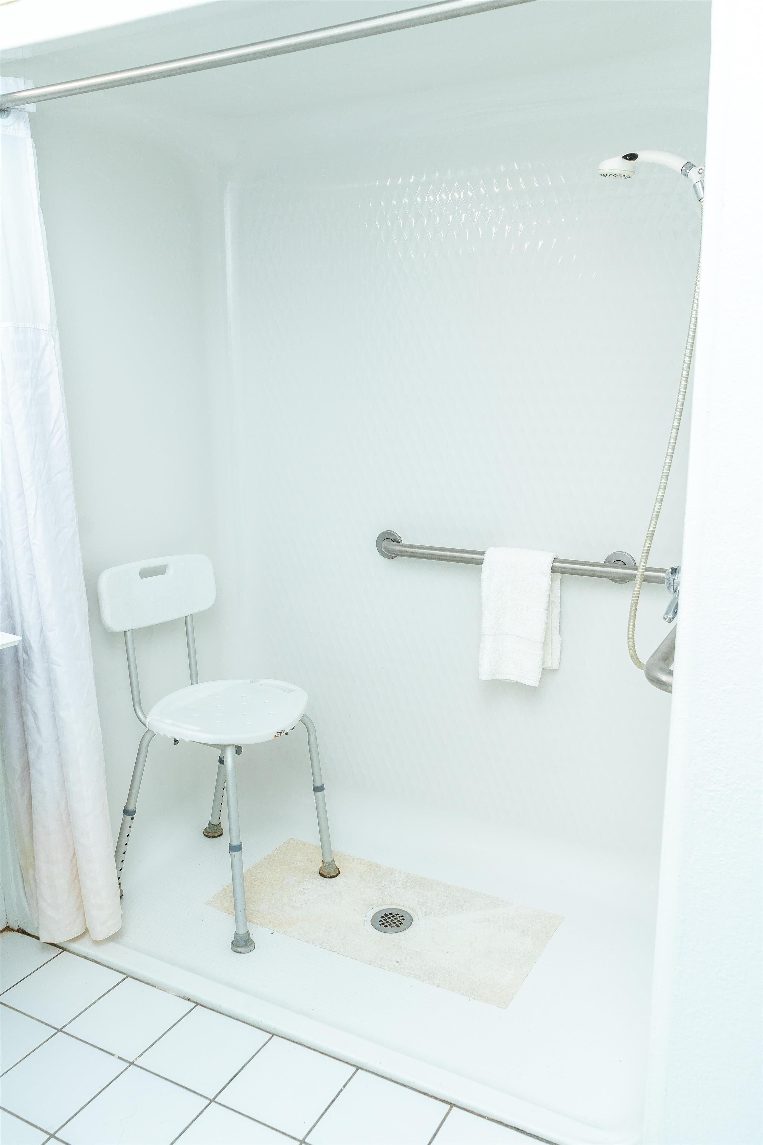 A white shower with a handheld showerhead, a grab bar, and a white chair.