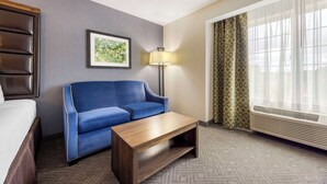 Ein Hotelzimmer mit einem Bett, einem blauen Sofa, einem Holztisch, einer Lampe, einem gerahmten Bild, einem Fenster mit Vorhängen und einer Klimaanlage.
