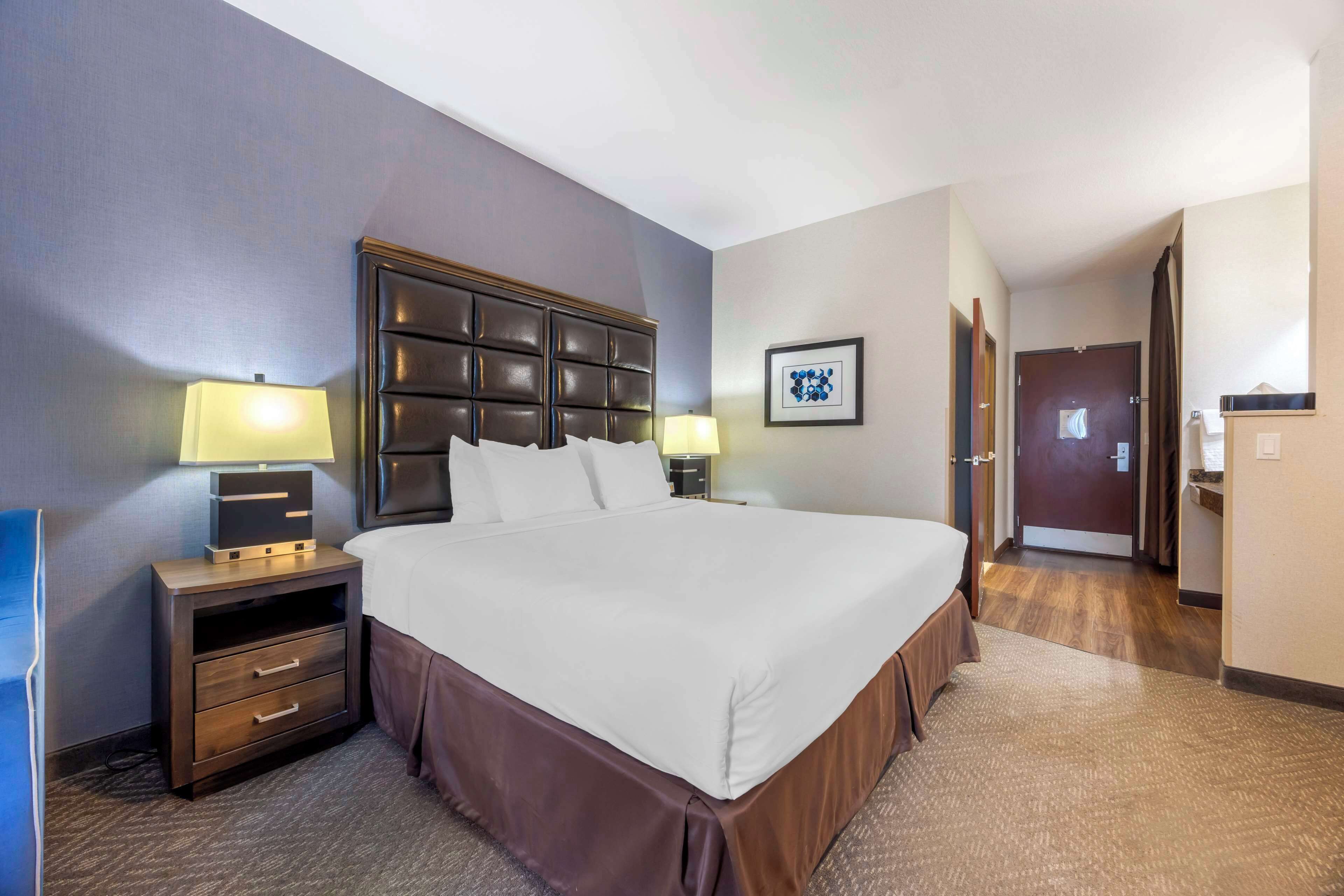 A hotel room with a large bed, a nightstand with a lamp, a dresser, and a door leading to another room.