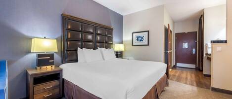 A hotel room with a large bed, a nightstand with a lamp, a dresser, and a door leading to another room.