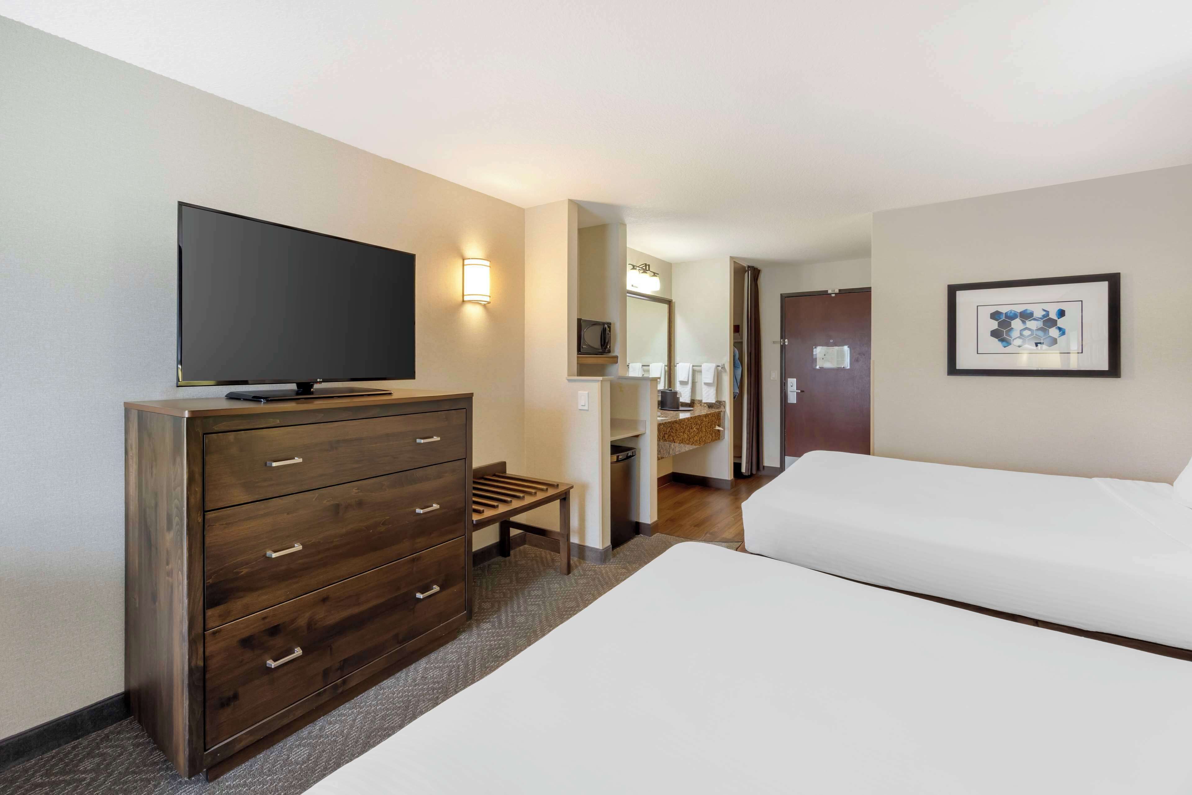 Habitación de hotel con televisión de pantalla plana sobre un tocador de madera, una cama, baño con espejo y un cuadro en la pared.