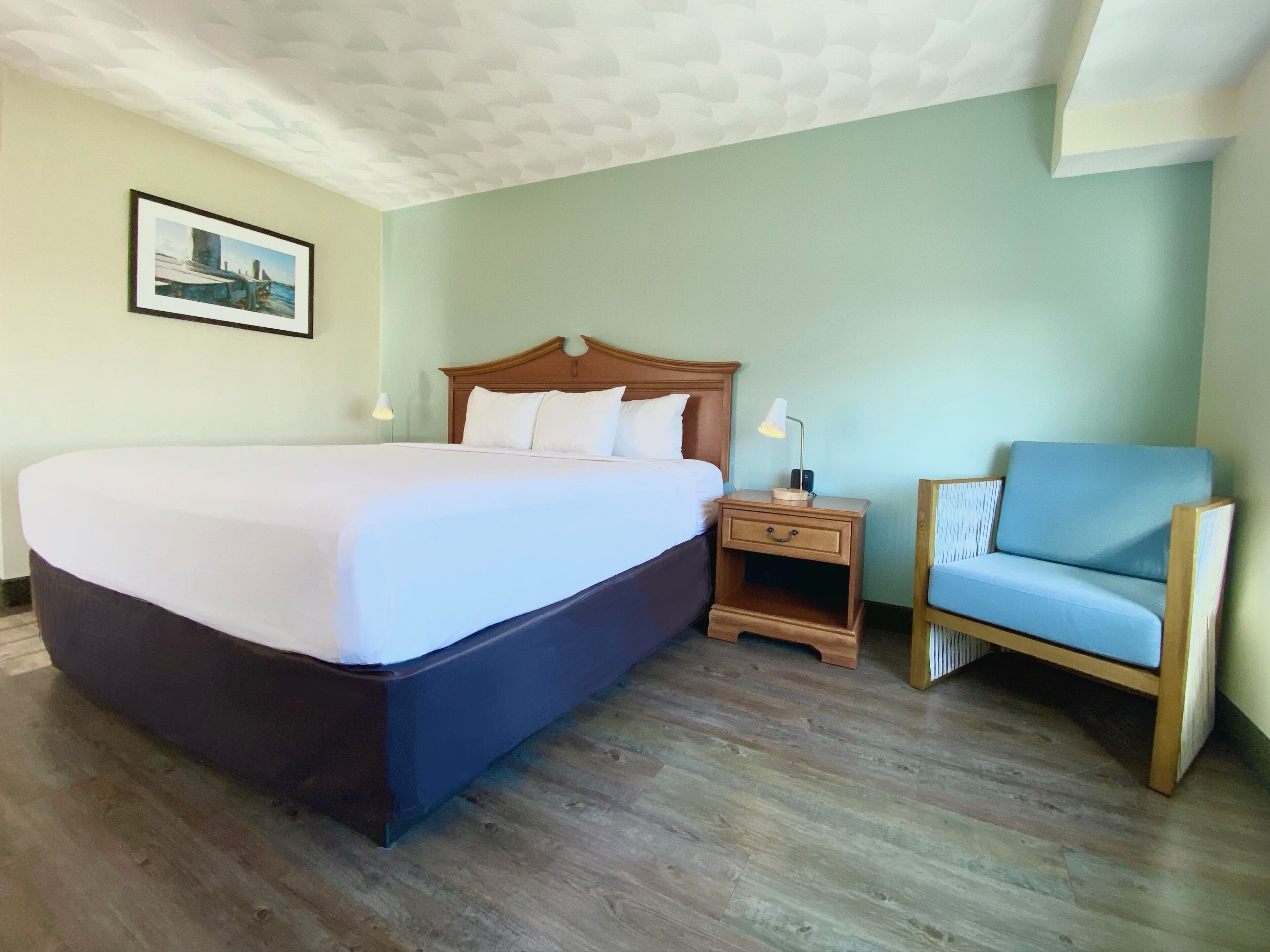 Una habitación de hotel con una cama grande, una mesita de noche de madera, una silla y un cuadro en la pared.