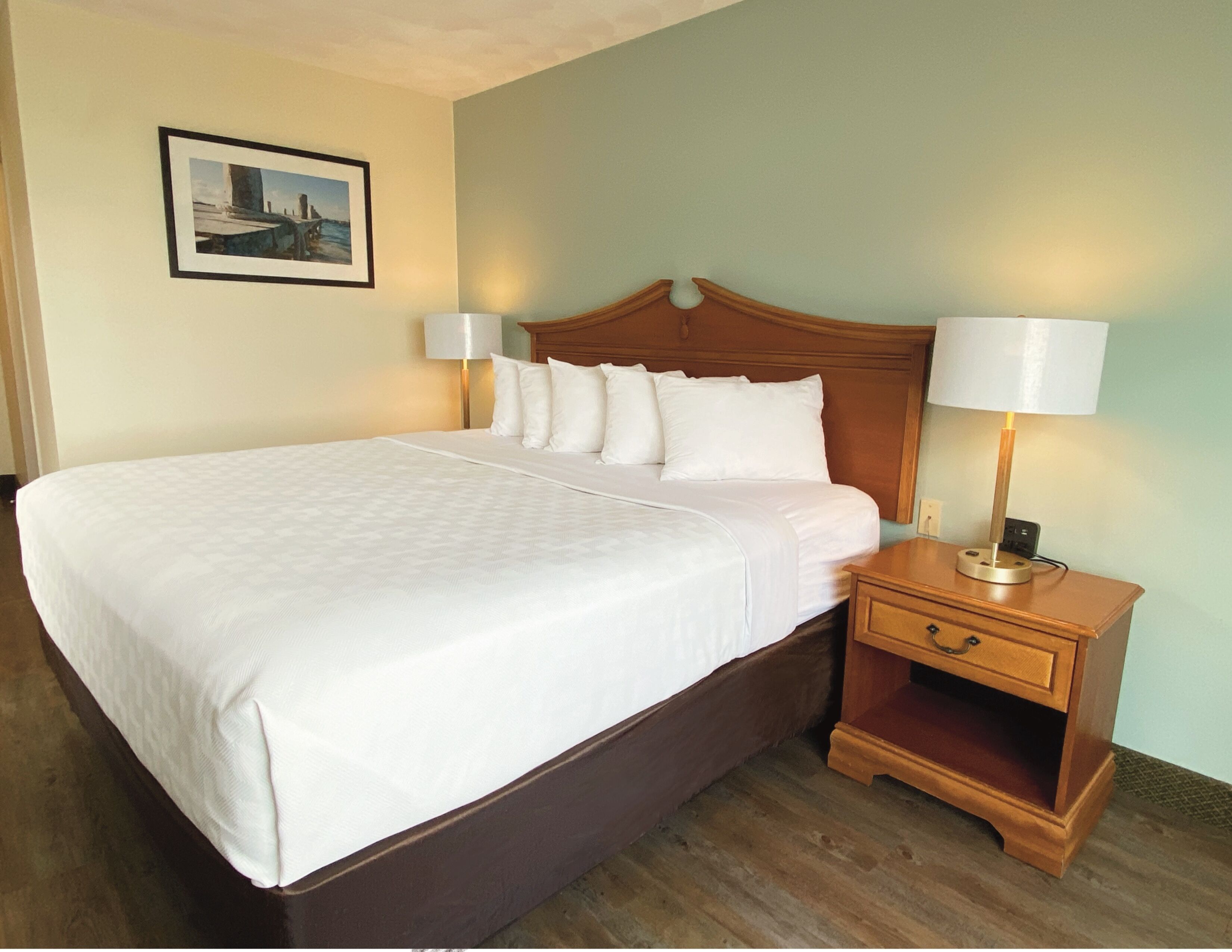 A neatly made bed with white linens and a wooden headboard, flanked by two bedside lamps and a small wooden nightstand with a drawer.