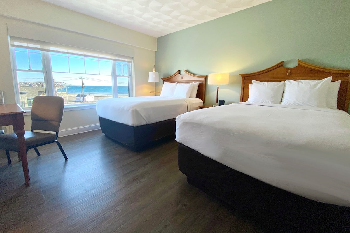 Een hotelkamer met twee bedden, een bureau en een stoel. Een groot raam biedt uitzicht op het strand.