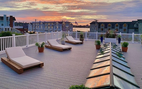 A rooftop terrace with lounge chairs and potted plants, overlooking a waterfront with buildings in the background.