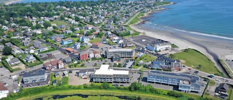 Vista aérea de un pueblo costero con zonas residenciales, una playa y un gran complejo de edificios con varios estacionamientos.