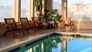 Une piscine intérieure à l’eau d’un bleu limpide, entourée de grandes fenêtres et de chaises en bois.