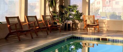 An indoor swimming pool with clear blue water, surrounded by large windows and wooden chairs.