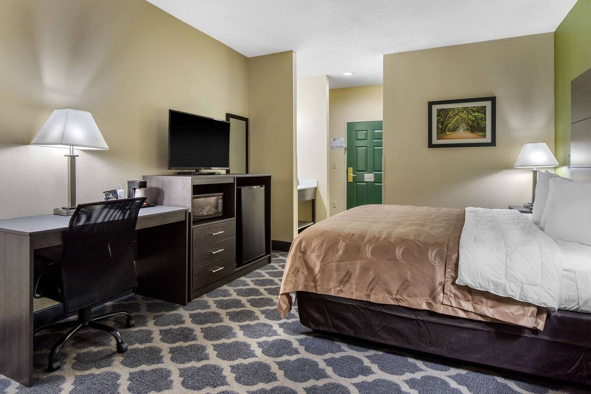 A hotel room with a bed, desk, chair, TV, and a door.