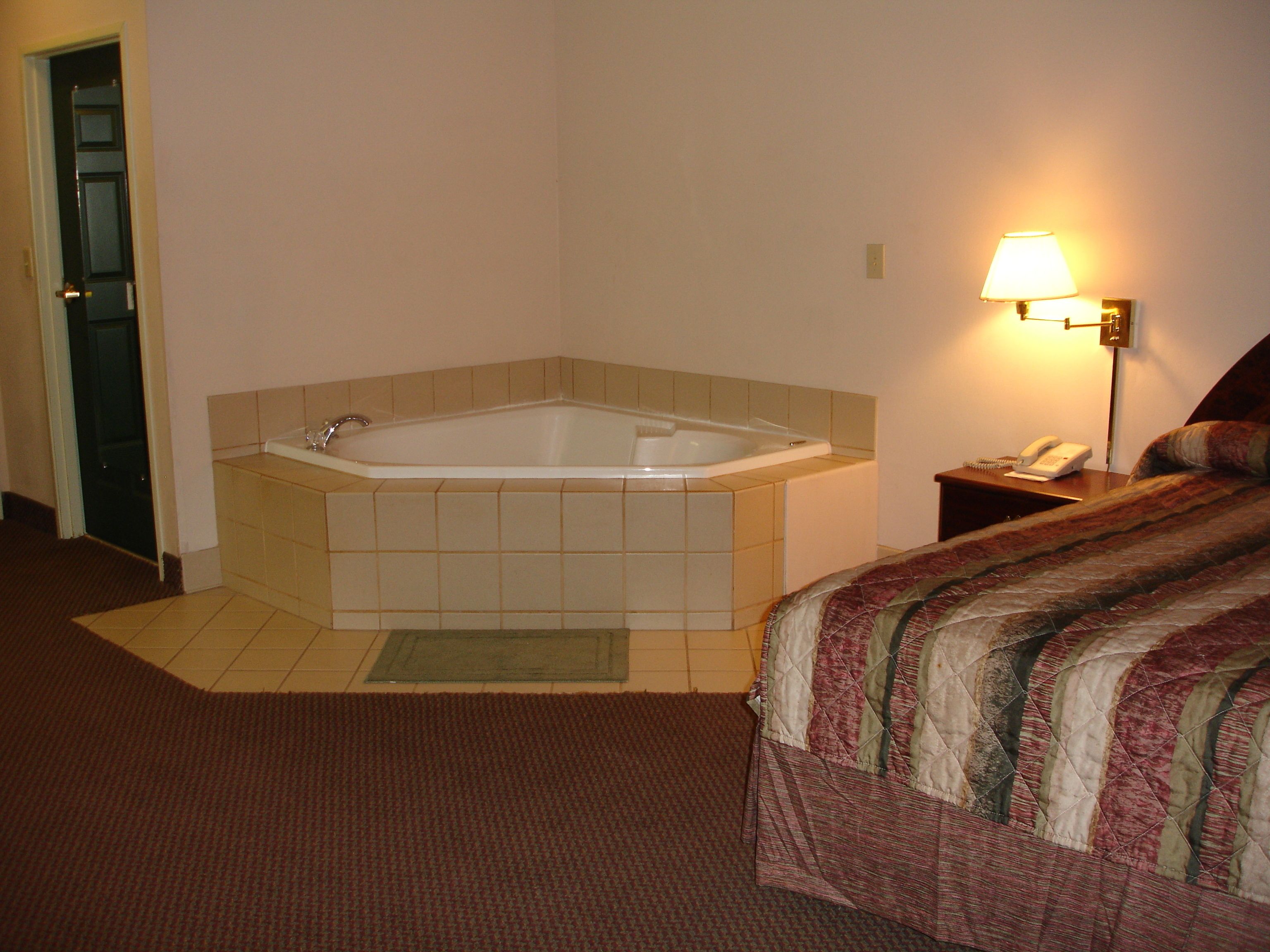 A hotel room with a corner bathtub, a bedside lamp, a telephone, and a patterned bedspread.