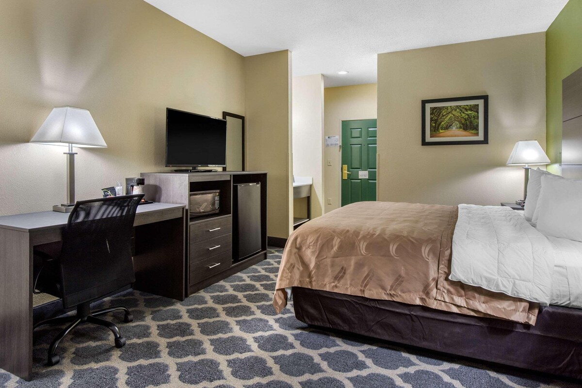 Habitación de hotel con cama, escritorio, silla, televisor y puerta.