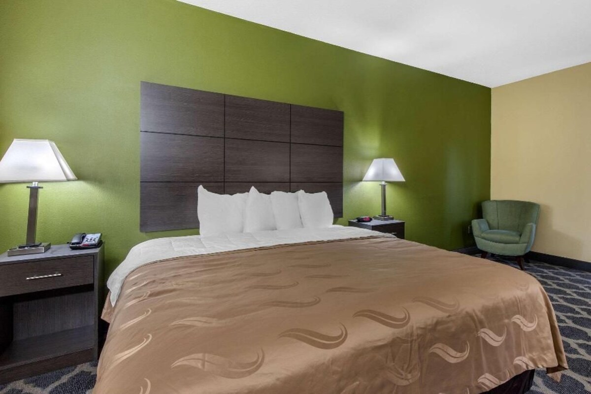 A hotel room with a large bed, two bedside tables, a green armchair, and a headboard with a decorative panel.