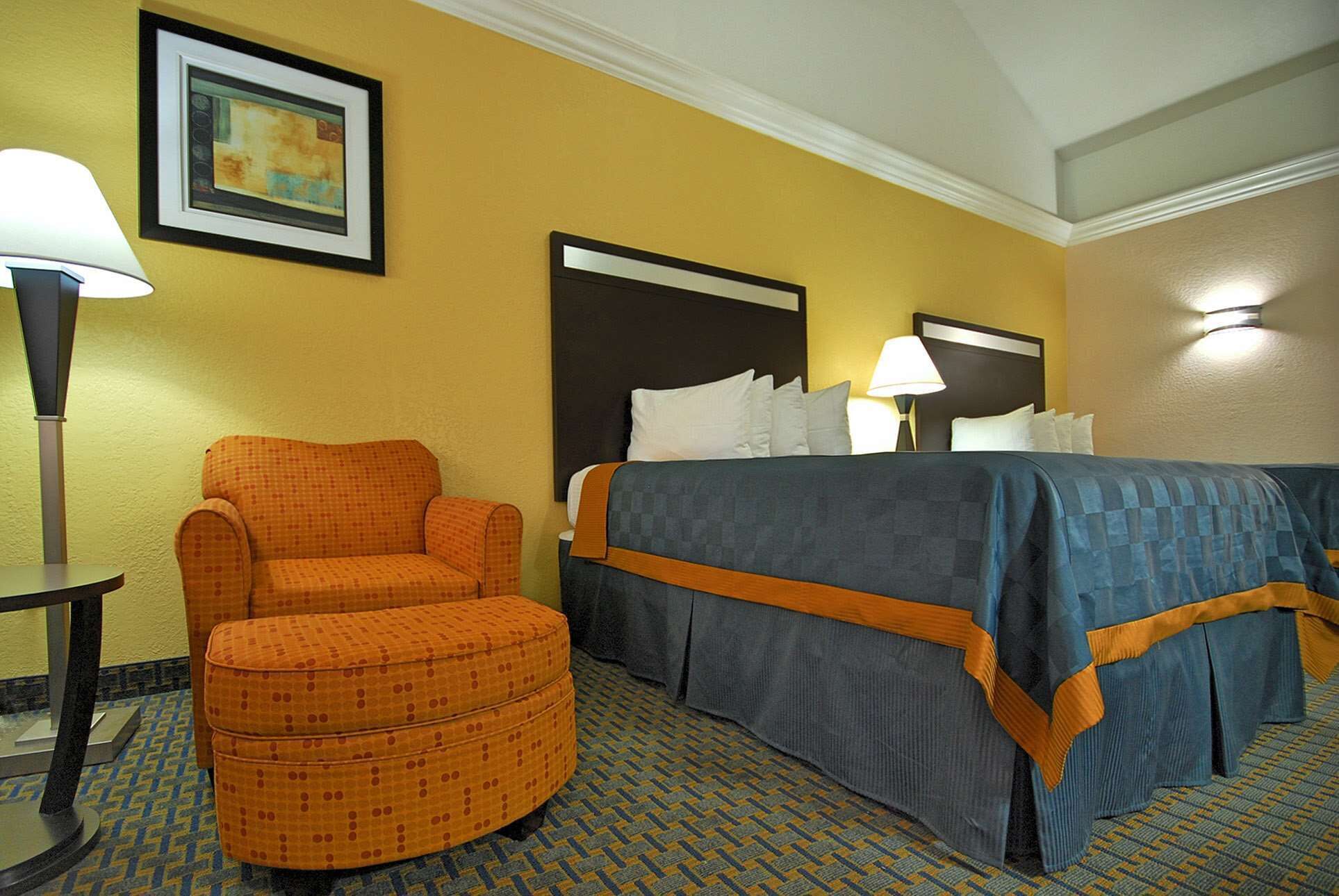 Una habitación de hotel con una cama, un sillón naranja, un pequeño reposapiés, un cuadro en la pared y una lámpara.