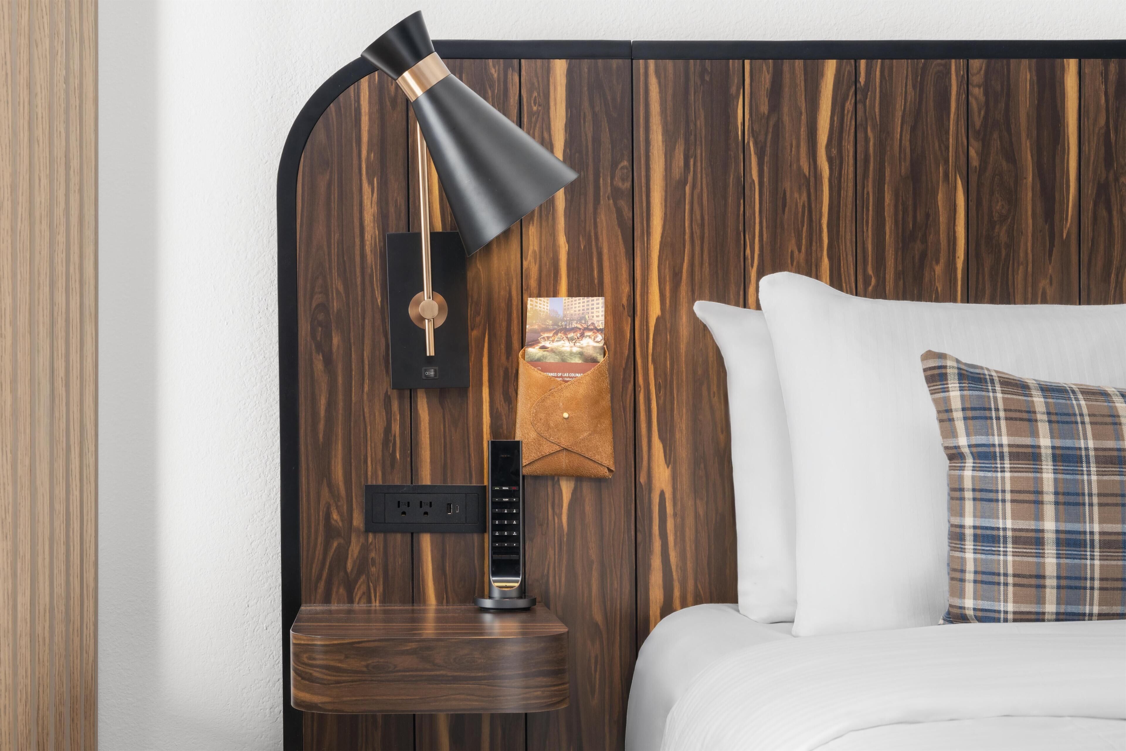 A neatly made bed with a plaid pillow, a wall-mounted lamp, a small wooden nightstand with a lamp, and a wall-mounted power outlet.